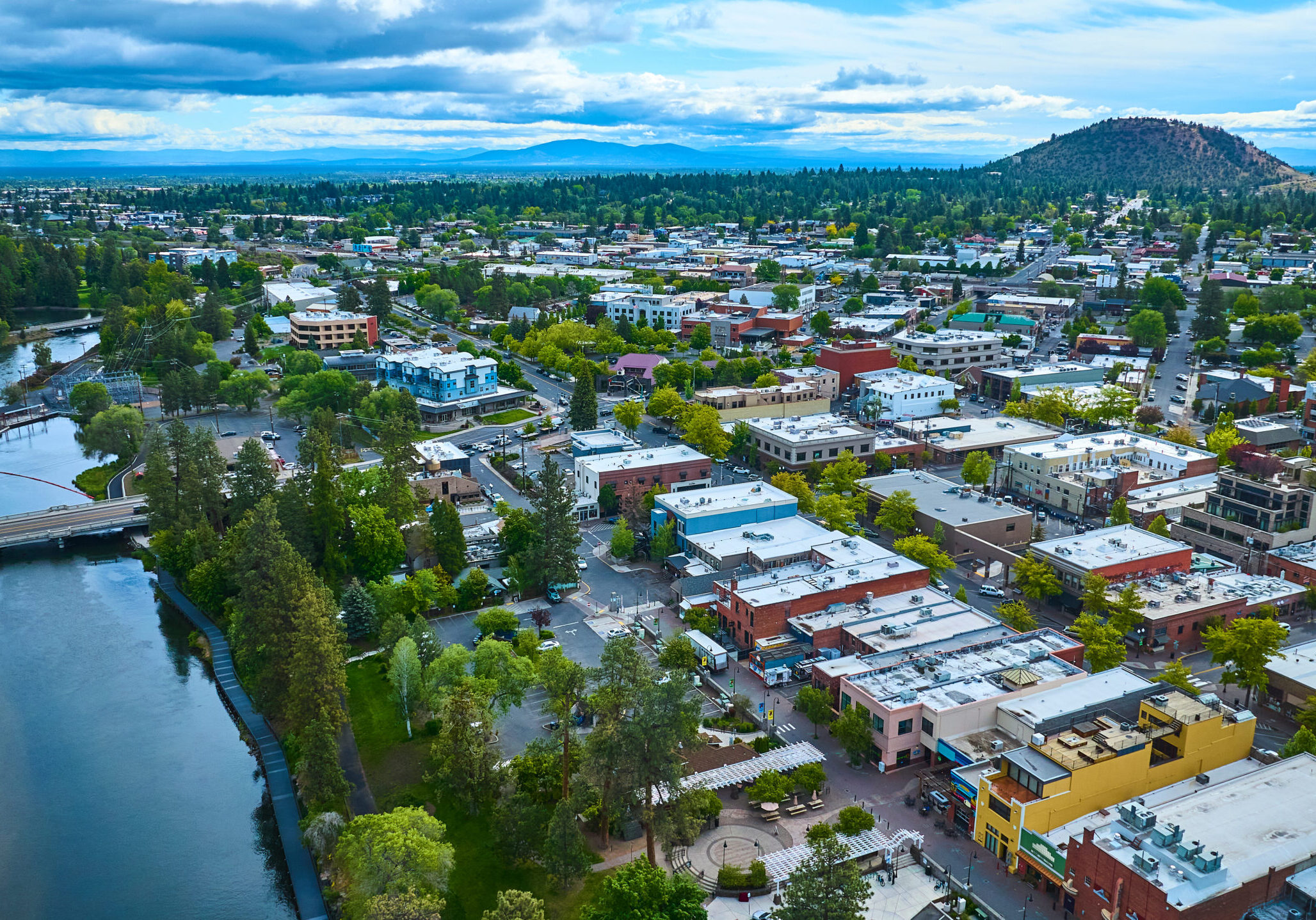 Aerial view of downtown Bend, Oregon. The vibrant town is nestled along a winding river, surrounded by lush greenery and a scenic hill. Perfect for promoting urban planning, tourism, or nature-integrated living.